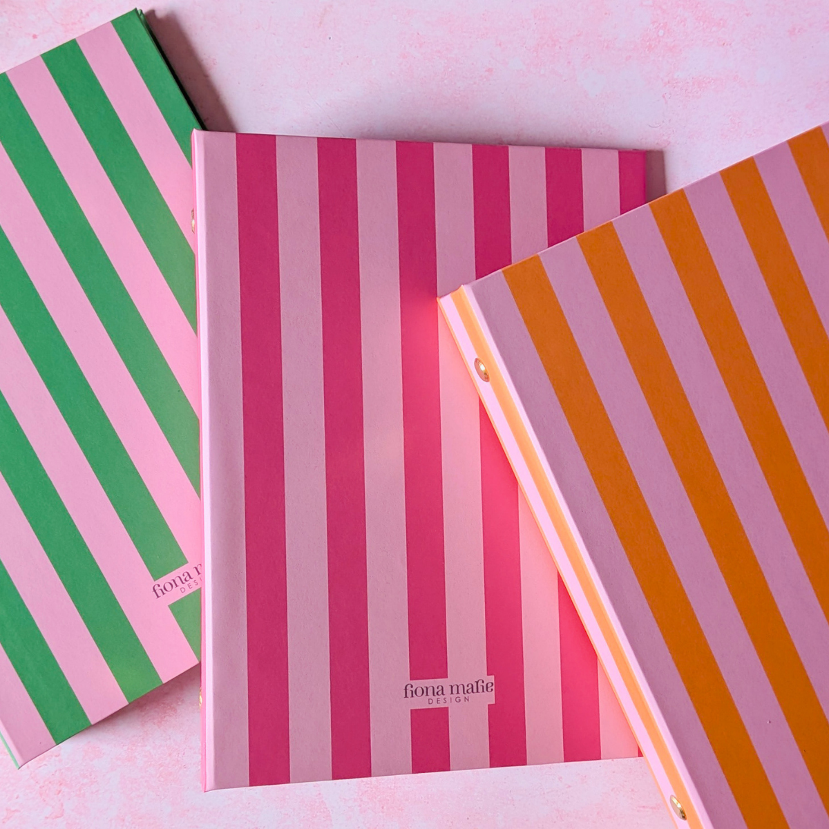 Three striped ring binders stacked diagonally on a light pink background. The top notebook has orange and lavender stripes, the middle has pink and white stripes, and the bottom has green and pink stripes.