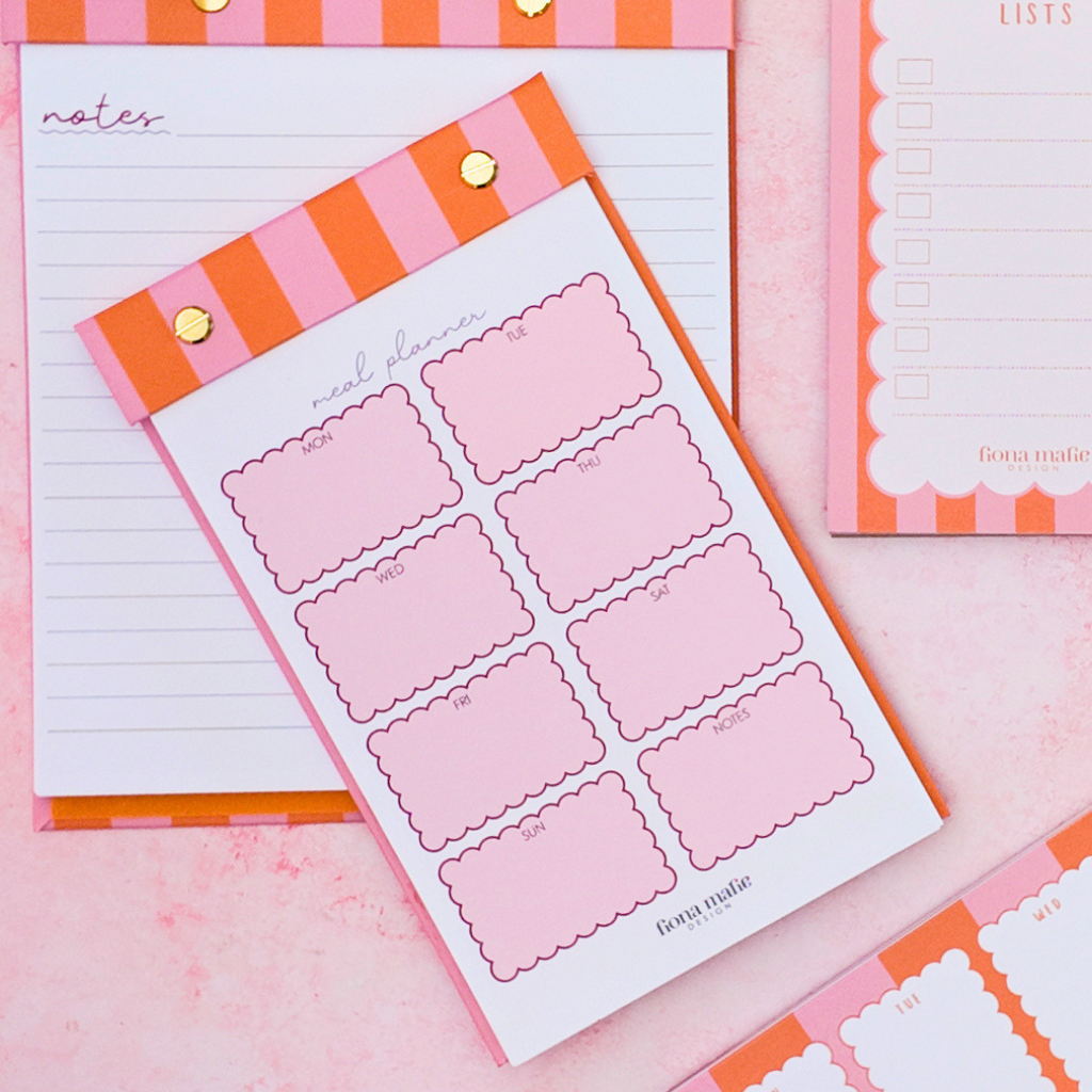 Pink and orange striped notepad and planner inserts arranged on a pink surface, with gold binder clips and a scalloped border on the pad edges.