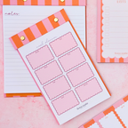 Pink and orange striped notepad and planner inserts arranged on a pink surface, with gold binder clips and a scalloped border on the pad edges.