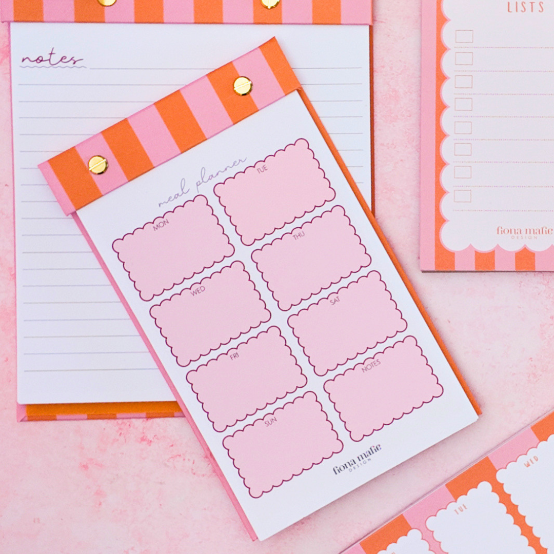 Pink and orange striped notepad and planner inserts arranged on a pink surface, with gold binder clips and a scalloped border on the pad edges.