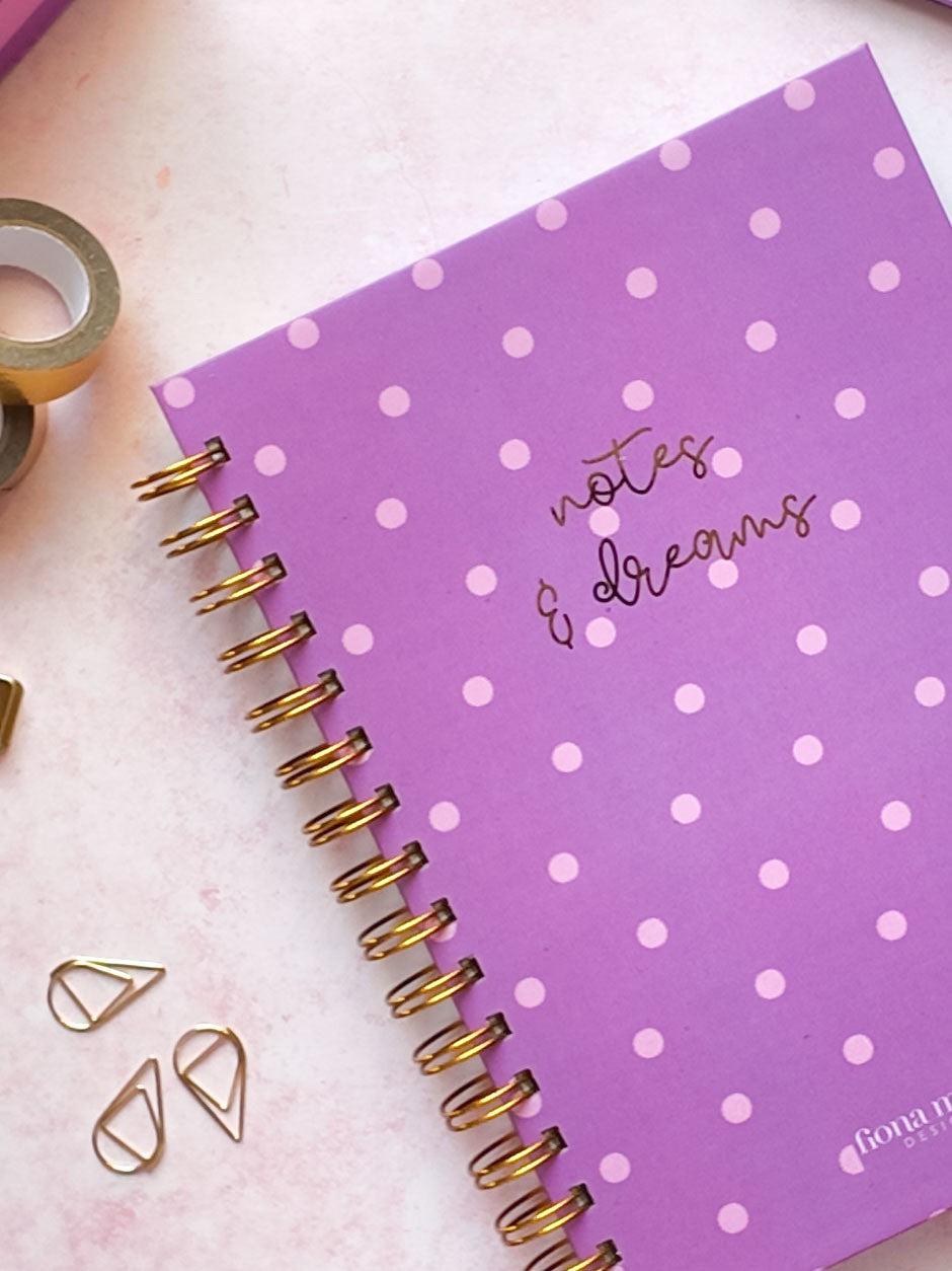 A purple spiral-bound notebook with pink polka dots and gold foil lettering that reads 'notes & dreams', styled on a light pink surface with gold tape and paperclip props.