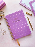 A purple spiral-bound notebook with pink polka dots and gold foil lettering that reads 'notes & dreams' is displayed on a pink surface with stationery props.