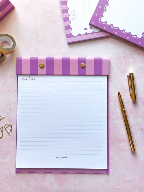 A pink and purple striped refillable notepad with gold heart-shaped clips, displayed on a pink background with gold stationery props including a pen, tape, and paperclips.