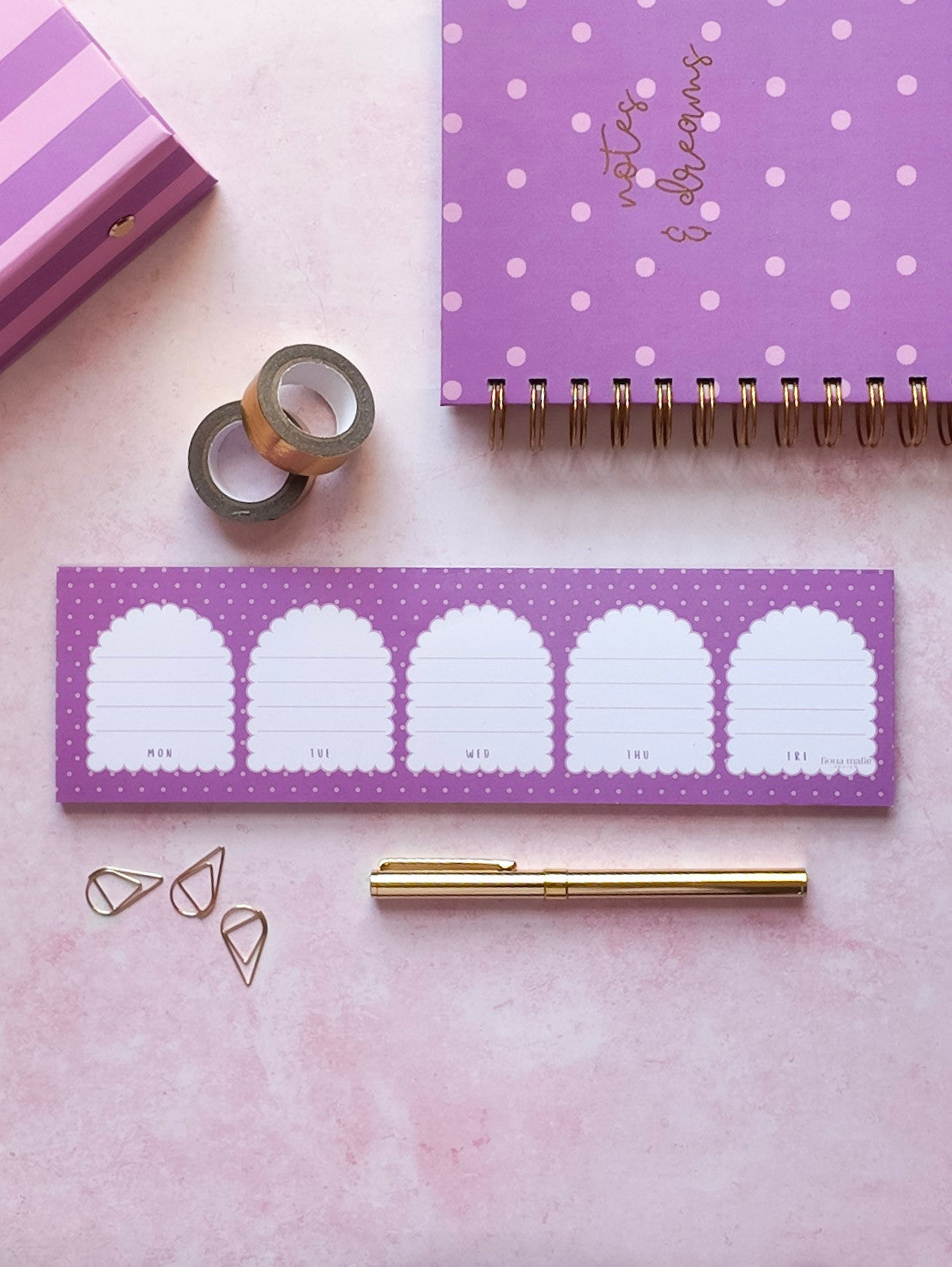 A styled flat-lay of a purple daily planner notepad with five arch-shaped lined writing sections, shown alongside a matching purple polka-dot spiral notebook, gold tape rolls, paper clips, a gold pen, and a pink striped box on a pink background.