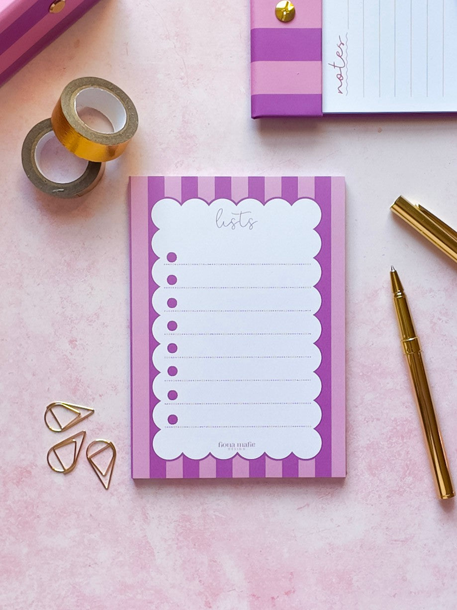 A pink and purple striped notepad with a white scalloped list page sits on a pink surface, styled with gold washi tape, gold paperclips, a gold pen, and another notepad in the background.