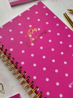A pink spiral-bound notebook with pink polka dots and gold foil text reading 'notes & dreams' on the cover, shown on a light surface with gold pens nearby.
