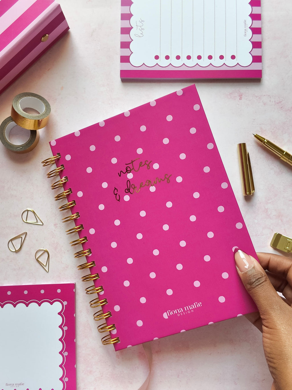 A pink spiral-bound notebook with pink polka dots and gold foil lettering is held by a hand over a pink surface, surrounded by stationery items including sticky notes, tape, paper clips, and a pen.
