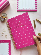 A pink spiral-bound notebook with pink polka dots and gold foil lettering is held by a hand over a pink surface, surrounded by stationery items including sticky notes, tape, paper clips, and a pen.