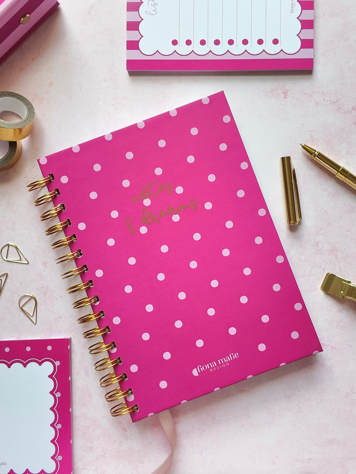 A pink spiral-bound notebook with pink polka dots and gold foil text reading 'hopes & dreams' on the cover, styled on a pale pink surface with gold and pink stationery accessories around it.