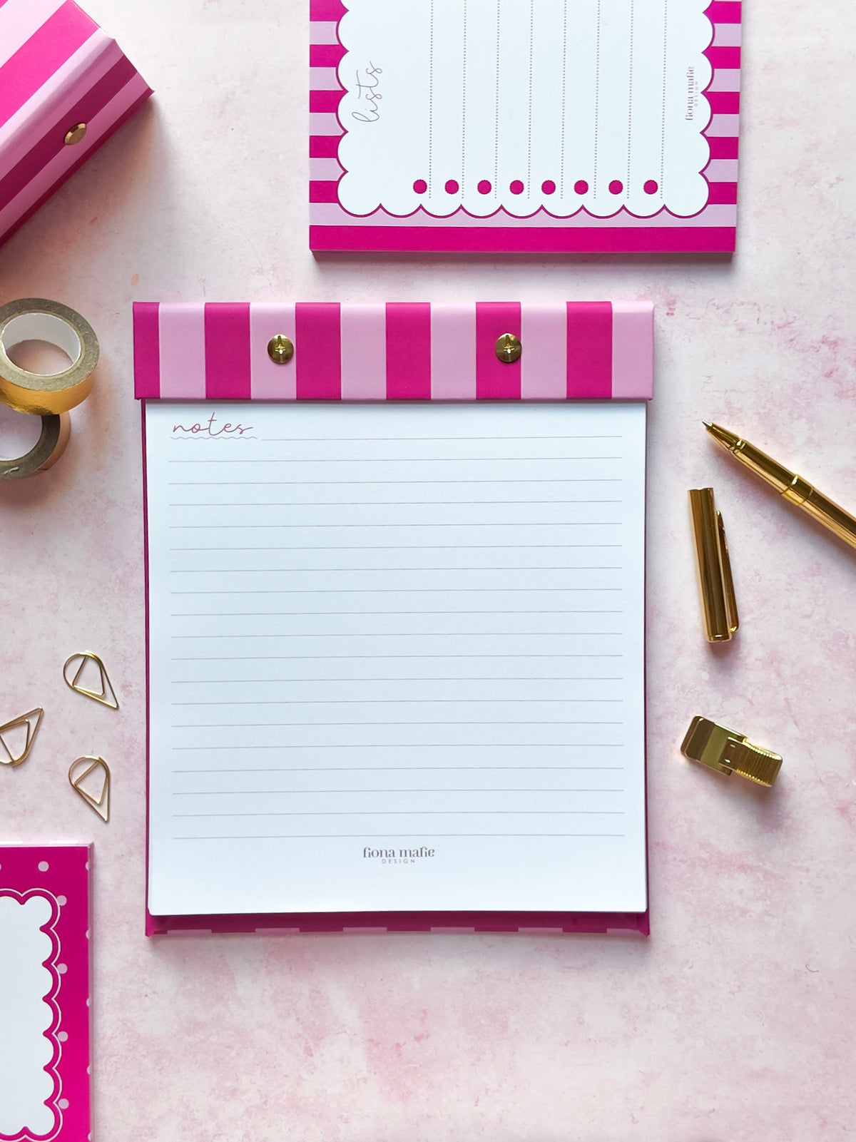 A pink and white striped refillable notepad with lined writing pages, styled in a flat lay on a pink surface with gold stationery accessories and a matching notepad at the top.