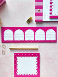 Pink and white stationery set featuring a long notepad with five arch-shaped lined writing areas, a square notepad with a scalloped border, a gold pen, and coordinating pink striped accessories on a pale pink background.