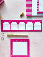 Pink and white stationery set featuring a long notepad with five arch-shaped lined writing areas, a square notepad with a scalloped border, a gold pen, and coordinating pink striped accessories on a pale pink background.