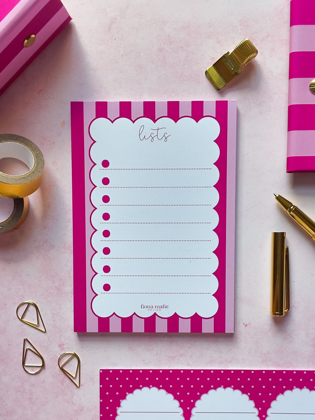 A pink and white striped notepad with a scalloped writing area labeled 'lists' is arranged on a pink surface with gold stationery props, including a pen, tape, clips, and additional pink patterned paper.