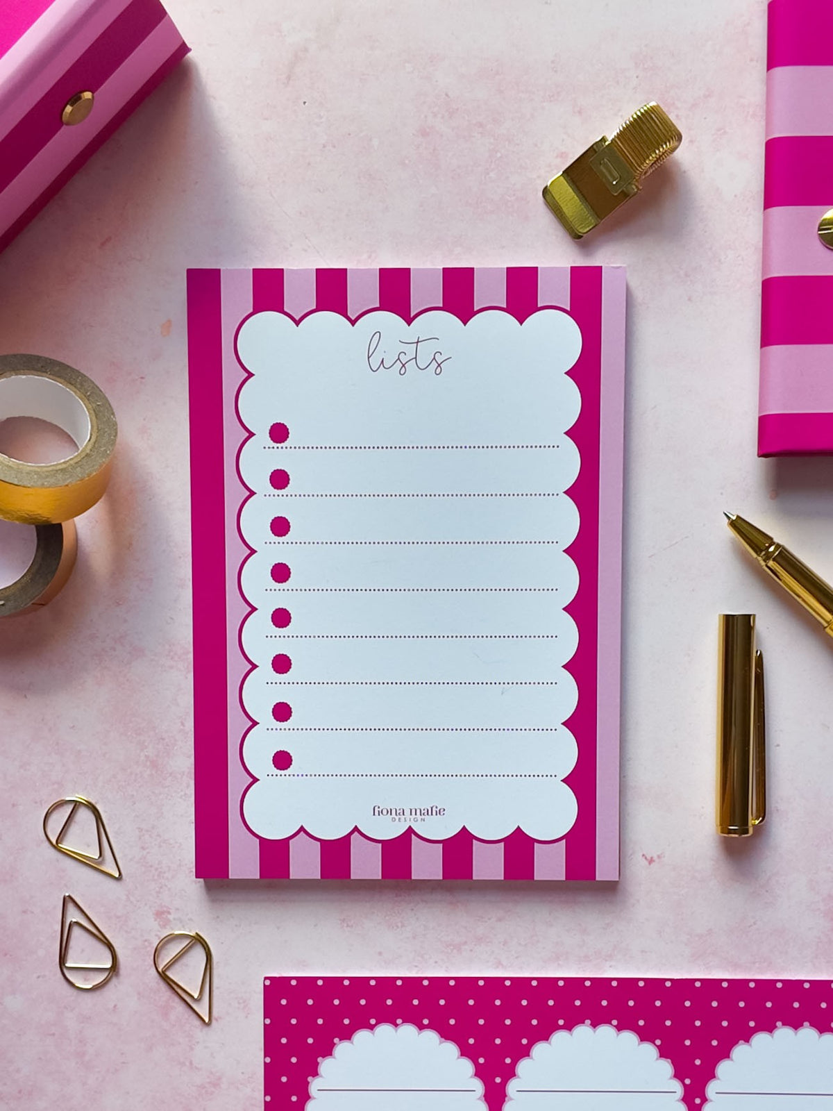 A pink and white striped notepad with a scalloped writing area labeled 'lists' is arranged on a pink surface with gold stationery props, including a pen, tape, clips, and additional pink patterned paper.