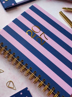 A spiral-bound 2027 dated diary with alternating pink and navy blue horizontal stripes, featuring gold foil '2027' on the cover and gold wire spiral binding, photographed on a light pink surface with gold and navy stationery props around it.