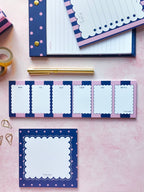 A styled flat-lay of pastel stationery items on a pink background, including a navy and pink striped notepad, a lined notebook, a gold pen, washi tape, paper clips, and a square memo pad.