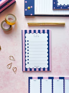 A pink and navy striped notepad with a scalloped 'lists' header is styled in a flat lay on a pink surface, surrounded by gold washi tape, gold paperclips, a gold pen, and matching stationery items.