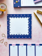 A styled flat-lay of a navy blue notepad with pink polka dots and a white scalloped center, surrounded by gold stationery accessories on a light pink background.