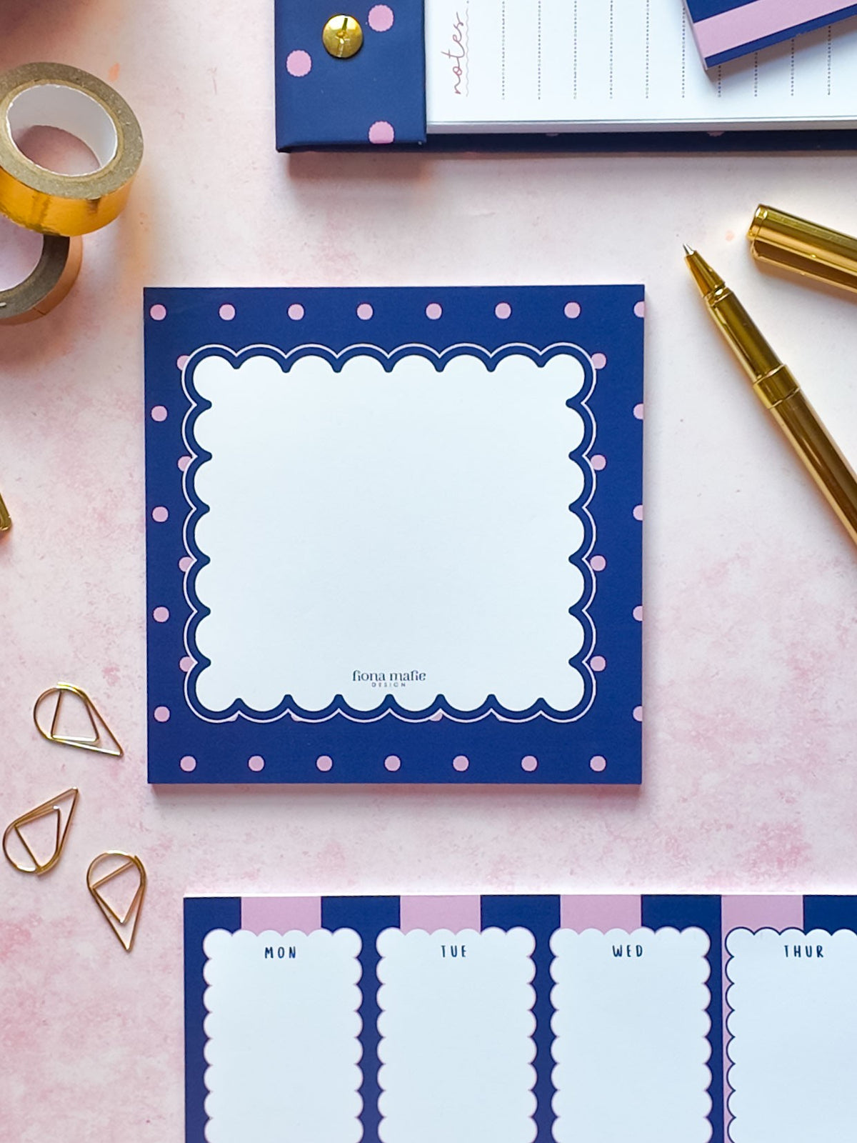 A styled flat-lay of a navy blue notepad with pink polka dots and a white scalloped center, surrounded by gold stationery accessories on a light pink background.