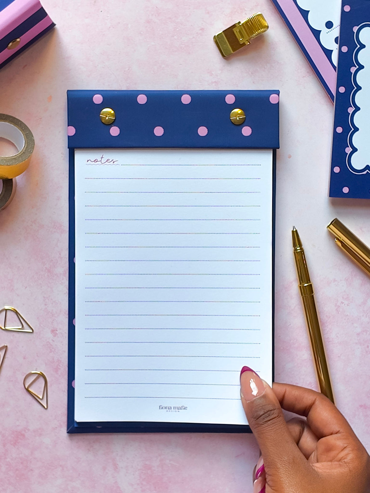 A navy blue refillable notepad holder with pink polka dots and gold snap buttons, shown on a pink surface. A hand is holding the bottom edge of the white lined notepad.