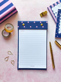 A navy blue lined refillable notepad with pink polka dots on the flap, styled in a flat lay on a pink background with gold and navy stationery accessories.