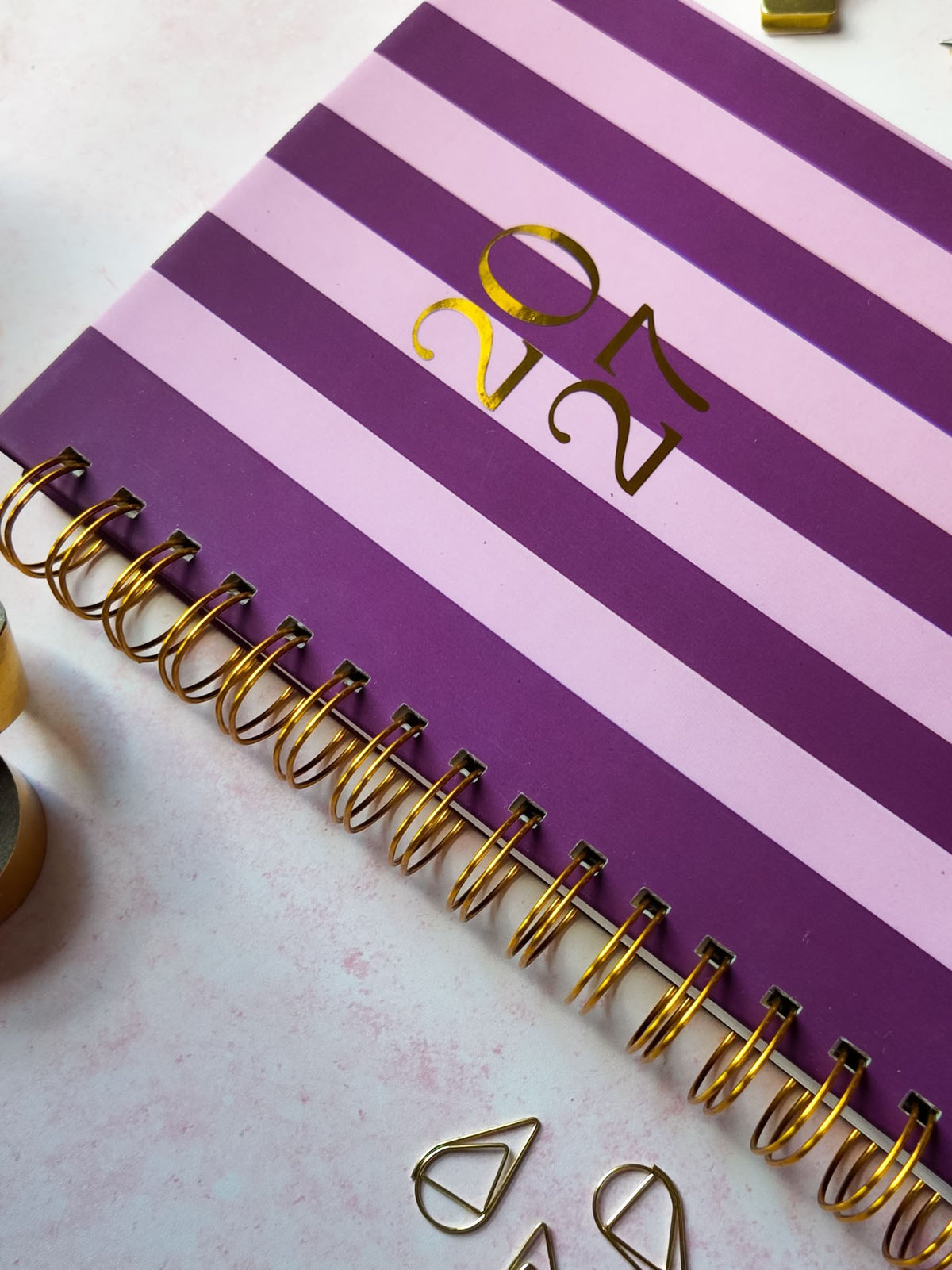 A spiral-bound 2027 dated diary with purple and light pink horizontal stripes, featuring gold foil lettering that reads 2027.