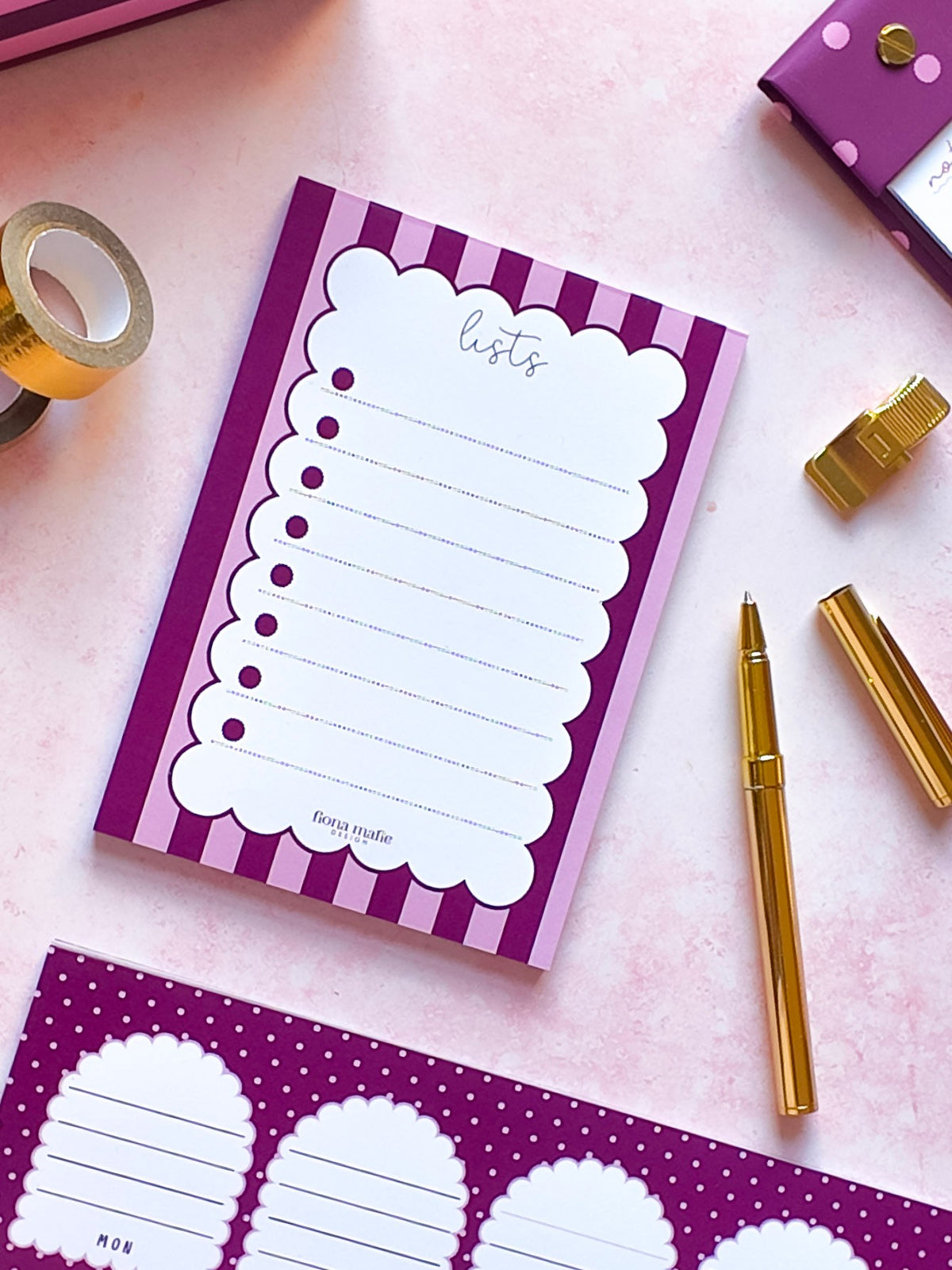 A purple and white striped notepad with a scalloped list page labeled 'lists' is arranged on a pink surface with gold stationery props, including a pen, tape, and matching paper sheets.