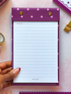 A hand with pink manicured nails holds a purple lined refillable notepad with pink polka dots and gold snap buttons. The notepad is shown on a pink desk surface with stationery props around it.