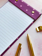 A purple lined refillable notepad with pink polka dots and gold snap buttons, styled on a pink surface with gold pens and a gold sharpener.