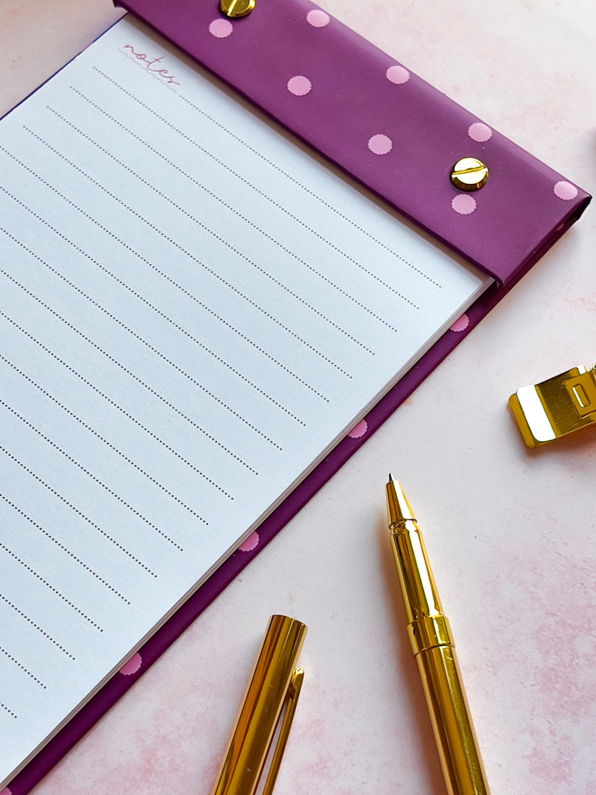 A purple lined refillable notepad with pink polka dots and gold snap buttons, styled on a pink surface with gold pens and a gold sharpener.