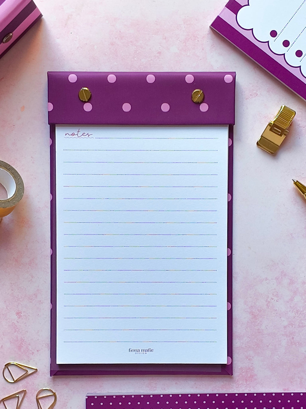 A purple and pink polka dot refillable notepad with a white lined writing page, styled in a flat-lay on a pink background with stationery accessories.