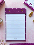 A purple and pink polka dot refillable notepad with a white lined writing page, styled in a flat-lay on a pink background with stationery accessories.