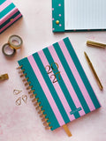 A pink and teal striped spiral-bound 2027 dated diary with gold foil numbers, styled on a pink surface with gold accessories and stationery.