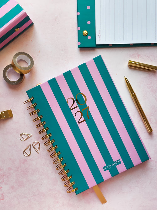 A pink and teal striped spiral-bound 2027 dated diary with gold foil numbers, styled on a pink surface with gold accessories and stationery.