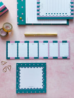 A styled flat-lay of teal, pink, and white stationery items on a light pink background, including a notepad, a weekly planner pad, washi tape, a gold pen, paper clips, and a small box.
