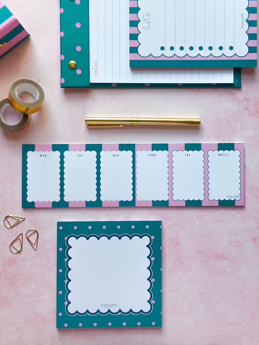 A styled flat-lay of teal, pink, and white stationery items on a light pink background, including a notepad, a weekly planner pad, washi tape, a gold pen, paper clips, and a small box.
