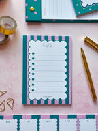 A teal and pink striped notepad with a scalloped white writing area labeled 'lists' is arranged on a pink surface with gold stationery props, including tape, paper clips, a pen, and additional notepads.