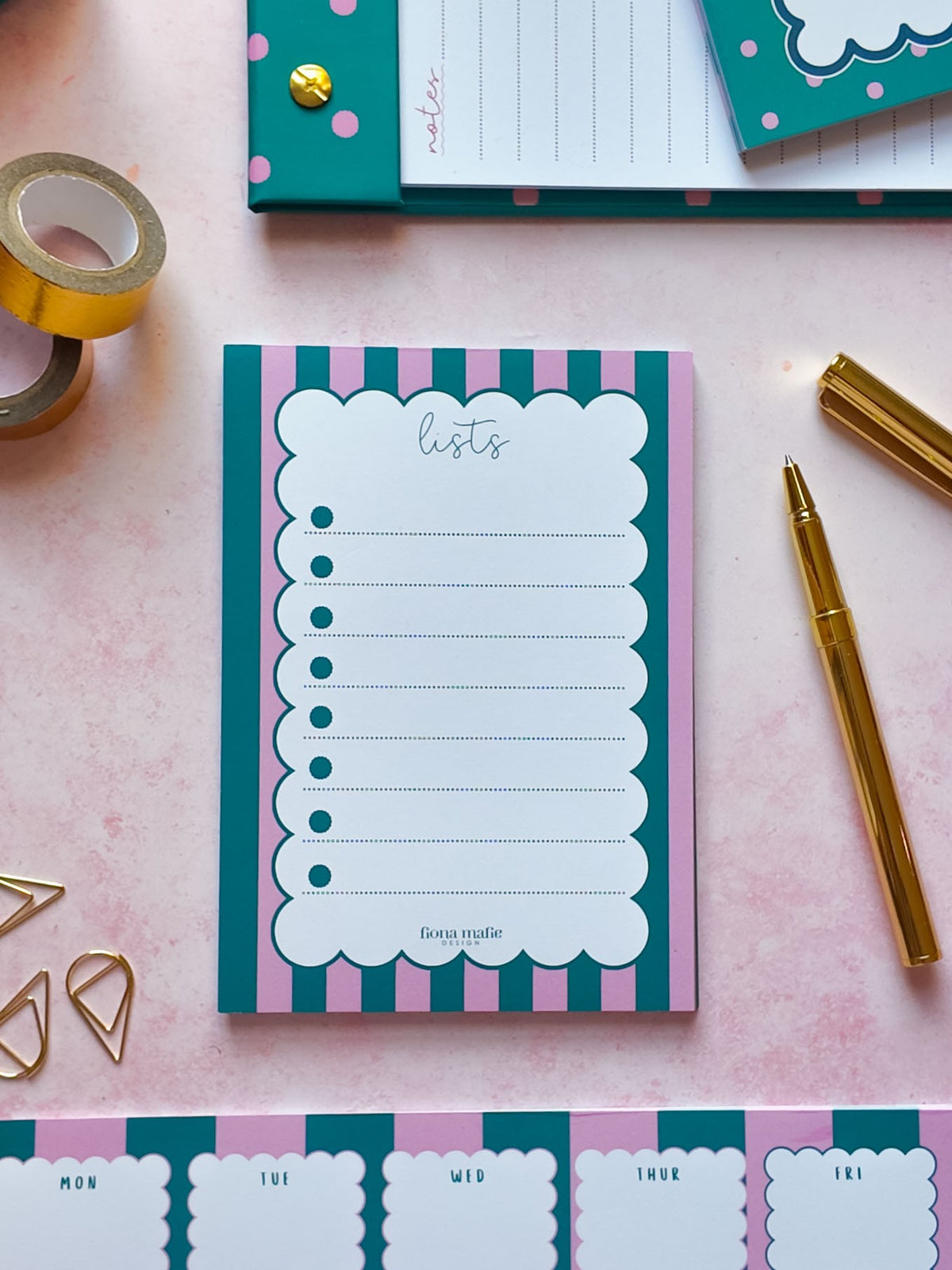 A teal and pink striped notepad with a scalloped white writing area labeled 'lists' is arranged on a pink surface with gold stationery props, including tape, paper clips, a pen, and additional notepads.