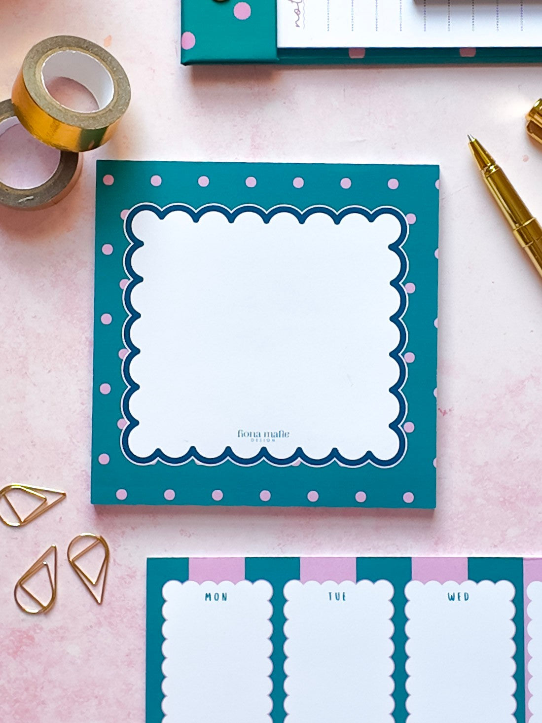 A flat-lay view of a teal notepad with pink polka dots and a white scalloped border, surrounded by gold stationery accessories on a pink background.
