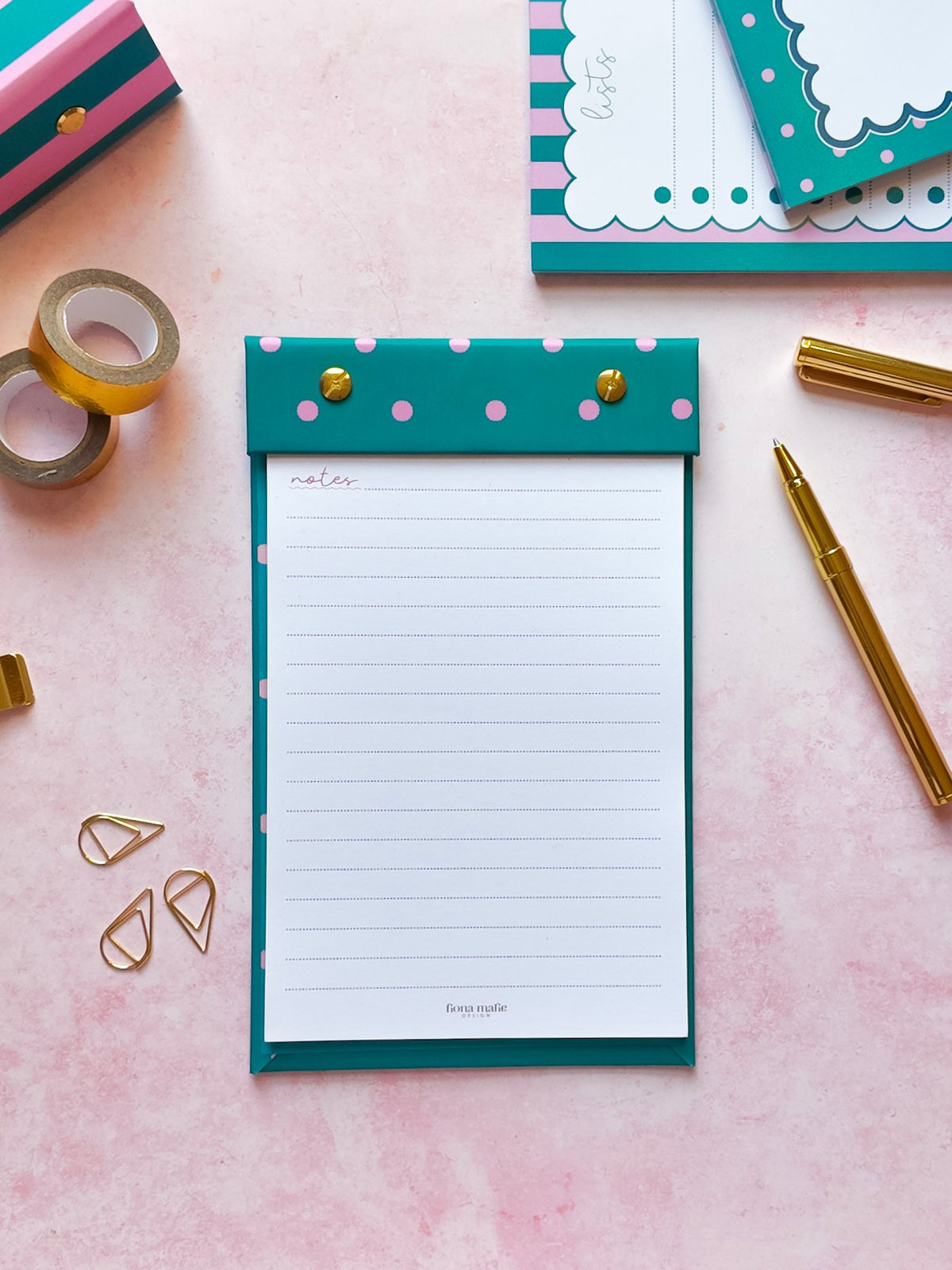 A teal and pink polka dot refillable notepad with gold snap buttons, displayed in a styled flat-lay on a pink background with gold stationery accessories.