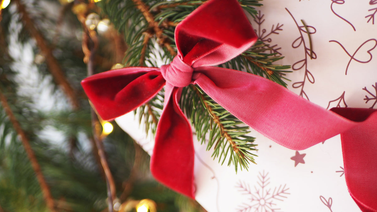 A close-up of a wrapped gift box with a large red velvet bow, decorated with green pine branches and printed with red snowflakes and stars on white paper. The background is a soft-focus Christmas tree with warm string lights.