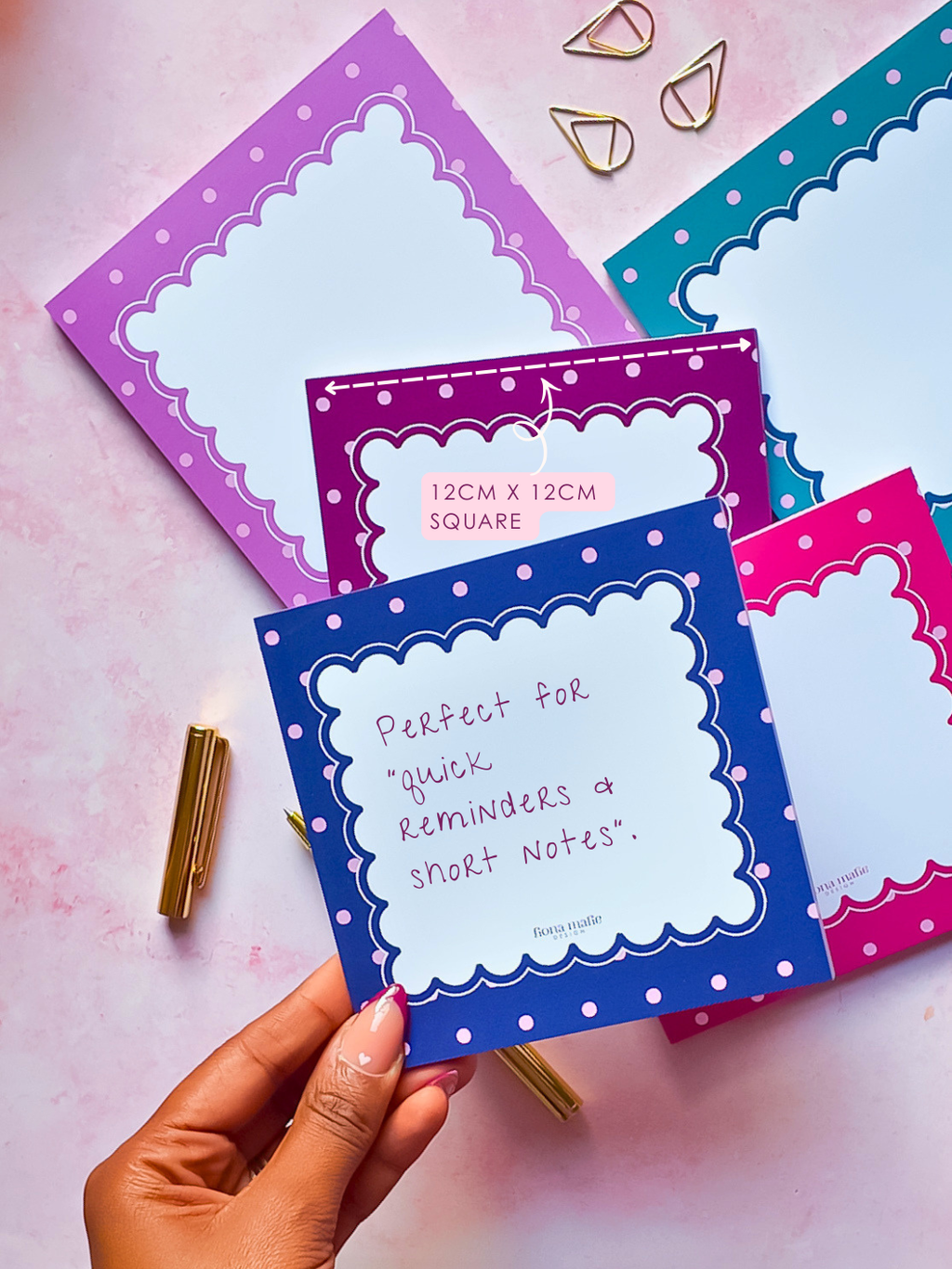 A hand holds a blue memopad with a white scalloped border and handwritten text, surrounded by other colorful notepads in pink, purple, and teal on a pink background with gold paperclips.