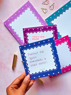 A hand holds a blue memopad with a white scalloped border and handwritten text, surrounded by other colorful notepads in pink, purple, and teal on a pink background with gold paperclips.