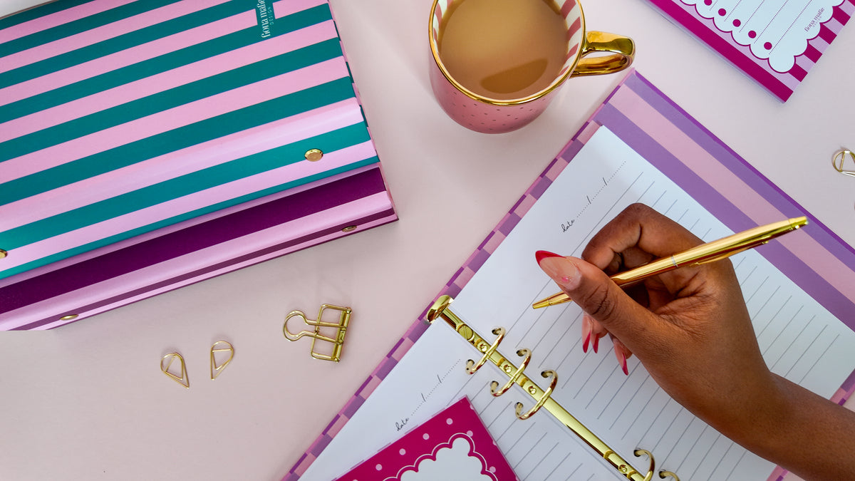 A styled desk scene with a striped ring Binder planner or notebook cover in pink, purple, and teal, an open ring binder notebook with lined pages, a gold pen being held by a hand, a pink mug with a gold rim, and small gold binder clips on a pale pink surface.
