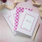 Three pink and white checkered notebooks with decorative designs on a pink surface.