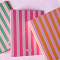 Three striped ring binders stacked diagonally on a light pink background. The top notebook has orange and lavender stripes, the middle has pink and white stripes, and the bottom has green and pink stripes.