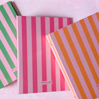 Three striped ring binders stacked diagonally on a light pink background. The top notebook has orange and lavender stripes, the middle has pink and white stripes, and the bottom has green and pink stripes.