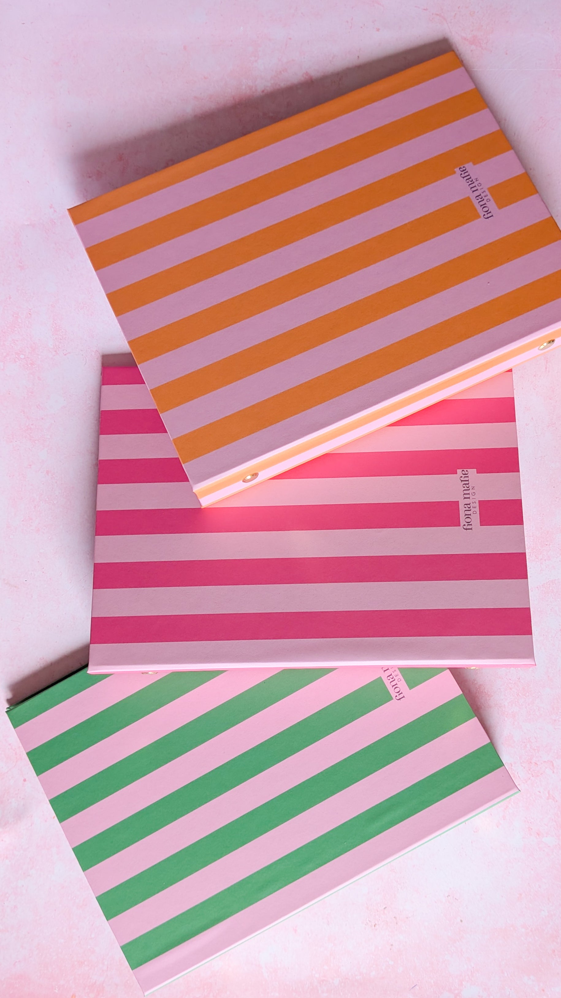 Three striped ring binders stacked diagonally on a light pink background. The top notebook has orange and lavender stripes, the middle has pink and white stripes, and the bottom has green and pink stripes.
