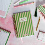 Notebook with green and pink striped cover on a white surface with stationery items.