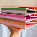 A hand holding a stack of colorful striped binder folders with gold metal rings, shown against a soft beige and light blue background.